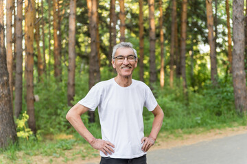 Naklejka na ściany i meble an elderly positive man of 60 with gray hair and a mustache on the background of a park in summer.portrait of a happy old pensioner in a white futblke smiling. a successful pension