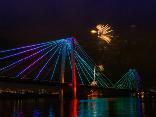 Obraz premium Blurred motion of bright flashes of fireworks in the night sky above the bridge. Fireworks in honor of Victory Day in Krasnoyarsk, Russia.