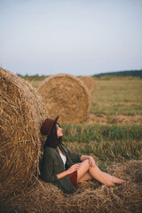 Beautiful stylish woman in hat sitting at hay bale in summer evening field. Happy young female...