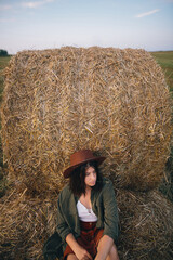 Naklejka na ściany i meble Beautiful stylish woman in hat sitting at hay bale in summer evening field. Happy young female relaxing at haystacks, summer vacation in countryside. Tranquility, countryside slow life