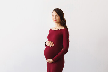 Young beautiful pregnant woman with long curly brunette hair, wearing burgundy dress, holding hands on her belly on white background.