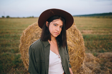 Beautiful stylish woman in hat standing at hay bale in summer evening field. Portrait of young fashionable female relaxing at haystacks, summer vacation in countryside. Tranquility
