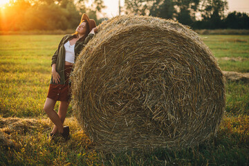 Beautiful stylish woman in hat relaxing on haystacks in sunset light in summer field. Atmospheric tranquil moment in countryside. Young female enjoying evening at hay bale in warm sunshine
