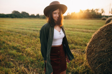 Obraz premium Beautiful stylish woman in hat walking at haystacks in sunset light in summer field. Atmospheric tranquil moment in countryside. Young female enjoying evening at hay bale in warm sunshine