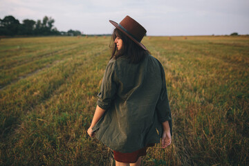 Naklejka na ściany i meble Beautiful carefree woman in hat walking and smiling in evening summer field. Young happy stylish female relaxing in countryside, enjoying evening. Atmospheric moment.