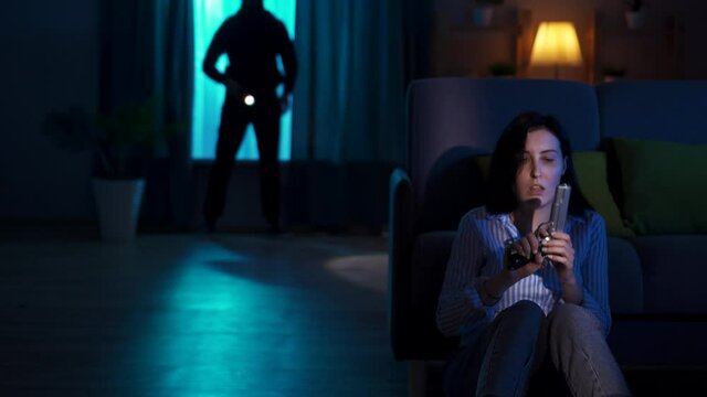 terrified young woman sitting on the floor with a gun in her hands and sees a robber with a fla in her house