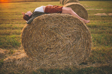 Beautiful carefree woman in hat lying on haystack in sunset light enjoying evening in summer field....