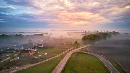 Aerial view of village, rural dirt road and trees covered by fog. Early misty morning sunrise panorama