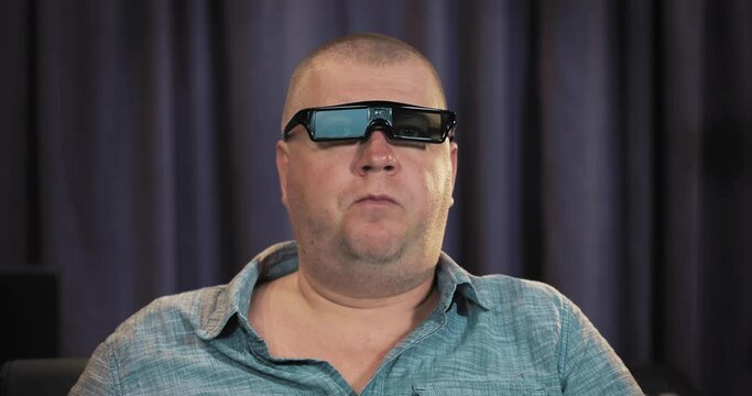 A man sits in a home theater in 3D glasses and eats popcorn. He's watching a movie on the screen. The man got tasteless, bad popcorn. He spat it back out. After that, the man continues to watch the mo