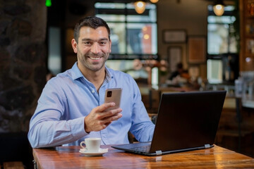 Hombre enviando un mensaje con el movil y su notebook laptop en un cafe despues del trabajo - obrazy, fototapety, plakaty