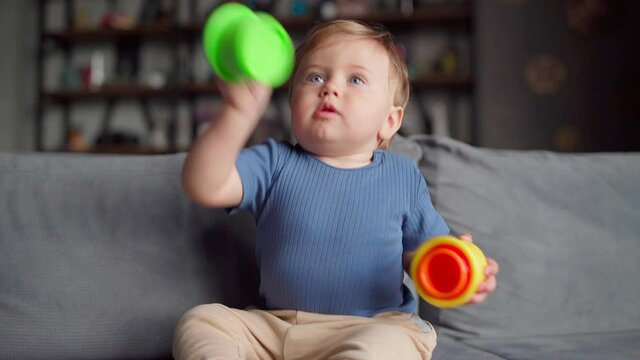 Adorable toddler having fun playing educational toy at home. Cute baby boy sitting on sofa and learning how to stack colorful plastic cups