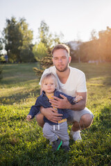Naklejka na ściany i meble father teaches his little son to walk, family walks in the park, dad holds the child's hands and helps to walk, evening walk, father-son relationship, trust in the family, beautiful baby