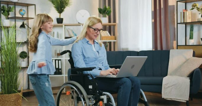 Pretty cheerful 10-aged girl running up to her beautiful skilled light-haired mother which sitting in wheelchair after accident and working on computer at home