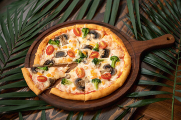 Pizza with broccoli, mushrooms and tomatoes. Vegan or vegetarian dish with vegetables on wooden cutting board among palm branches. Italian dinner or lunch. Fast food. Horizontal format.