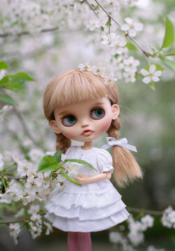 Blonde doll blythe with bangs in a white dress sits on a twig in a spring cherry orchard among white flowers
