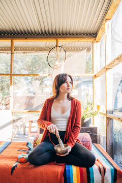 Woman meditating and playing brass singing bowl looking out window at sunlight