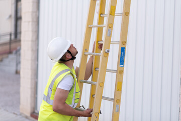 telecommunications technician in safety helmet and reflective vest, deploying a yellow ladder while looking upward. - obrazy, fototapety, plakaty