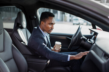 Fototapeta na wymiar Side view of focused african man sitting on driver's seat with cup of coffee and using dashboard for navigation. Businessman in formal wear taking stop for checking direction.