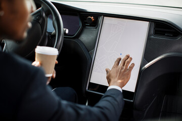 View from shoulder of african businessman in suit using electronic dashboard while sitting inside luxury electric car. Concept of people, modern technology and ecological vehicle.