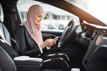 Side view of pretty arabian woman in hijab sitting on driver's seat and using modern smartphone. Business lady checking work emails on mobile inside luxury car.