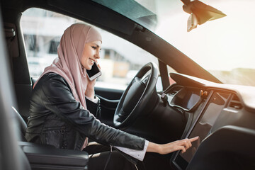 Side view of muslim woman in hijab tapping on electronic dashboard of luxury car while having mobile conversation. Concept of modern technology, people and transport.