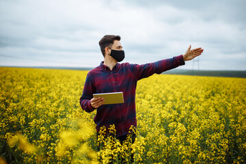 A successful farmer shows the rapeseed crop. A young agronomist in a black protective mask checks a rapeseed field with a tablet in his hand.