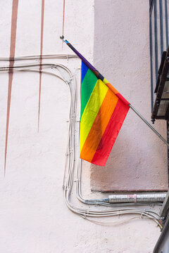 Pride flag hanging on a wall with a cream textured background, a balcony to the right side and some painted spikes falling in the background