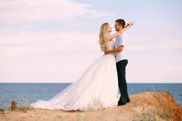Fototapeta na wymiar Beautiful bride and groom hug on the background of the sea, standing on the edge of the mountain. The bride is dressed in an elegant wedding dress and looks passionately at her beloved. Newlyweds hug