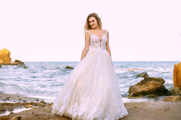 Fototapeta na wymiar A beautiful bride in a white wedding dress stands on the shore of the ocean and sandy rocks on the wedding day. Elegant young bride enjoying her wedding day posing for a photographer