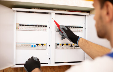 Confident young electrician in helmet and gloves is checking with an electrical panel with fuses