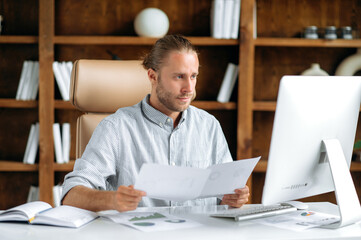 Smart focused young adult caucasian man, sits at the work desk in office, in stylish clothes, uses a computer, looks through documents and charts, thinks over financial strategy, working concentrated