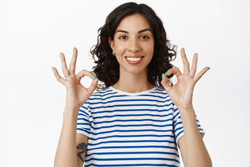 Portrait of smiling girl shows okay OK signs, look satisfied, recommend good company, perfect quality, praise good job, well done, standing pleased against white background