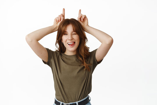 Cheeky young woman winking, showing tongue and smiling, making bull horns gesture above head, standing funny and playful against white background