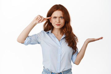 Frustrated redhead woman pointing at head and shrugging, scolding someone stupid, confused with...