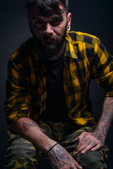 Fototapeta premium Tattooed man with a beard wearing checkered shirt is posing seated on a chair in a studio