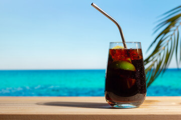 Glass of Cuba Libre cocktail on tropical beach, blue sea on background. Summer resort concept, alcohol drink. Rum cola cocktail. 