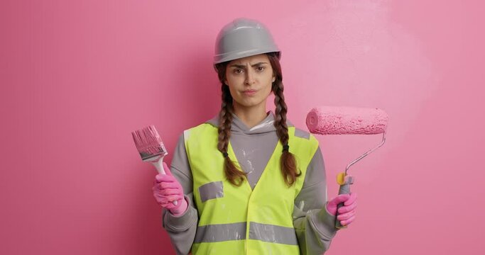 Hesitant unaware woman builder in working uniform looks clueless at painting roller and brush chooses tool for refurbishment busy doing house repair isolated over pink background. Industry concept