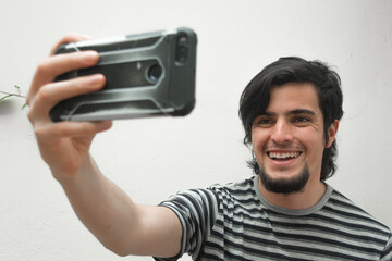Portrait of a young man with beard holding a phone on his hand smiling and taking a selfie. Model wearing a black and white striped t-shirt. Happiness and confidence face or facial expressions.