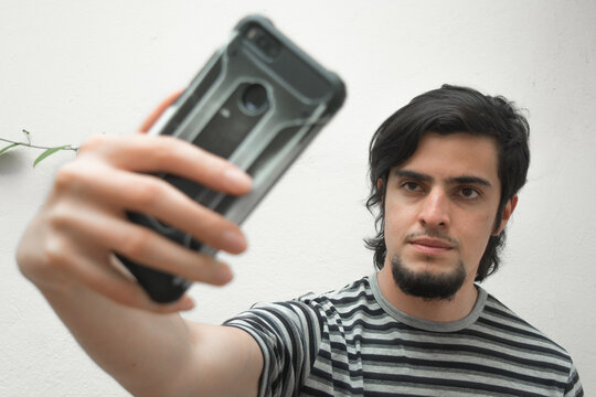 Portrait of a young man with beard holding a phone on his hand taking a selfie with serious or handsome expression. Model wearing a black and white striped t-shirt. 