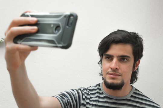 Portrait of a young man with beard holding a phone on his hand taking a selfie with serious or handsome expression. Model wearing a black and white striped t-shirt. 
