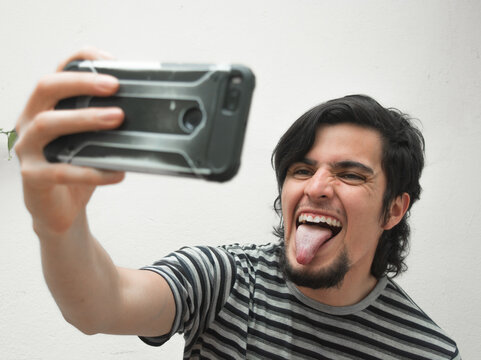Portrait of a young man with beard holding a phone on his hand smiling tongue out taking a selfie. Model wearing a black and white striped t-shirt. Happiness and confidence face or facial expressions.