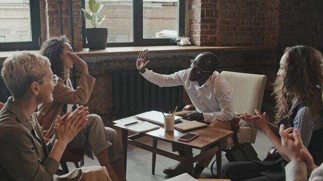 Dollying-out slowmo of diverse group of young male and female business people celebrating success of important project clapping hands and giving high-fives to each other in modern coworking office