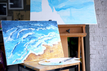 Acrylic sea and beach painting on drawing stand with palette and brush. Blue wave on golden sand hand painting with brush and knife palette technique. Art on canvas.
