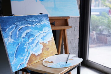 Acrylic sea and beach painting on drawing stand with palette and brush. Blue wave on golden sand hand painting with brush and knife palette technique. Art on canvas.