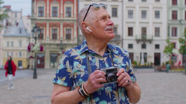 Senior man tourist with retro photo camera, smiling, listening music on earphones, dancing on summer city street center of Lviv, Ukraine. Photography, travelling vacation. Active life after retirement