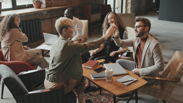 Slowmo of cheerful coworking team giving high-fives to each other celebrating success of project while their multi-ethnic colleagues discussing business in background at cozy loft-style workspace