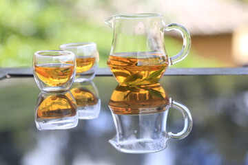 Hot clear tea in transparent jag and glass on table with reflection in nature. Tea set in tea field at Mae Hong Son, Thailand.