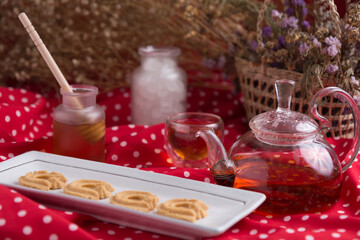 Tea pot and tea cup on wood table with honey and suagr in mag with flower basket on red cloth napkin, with butter cookies