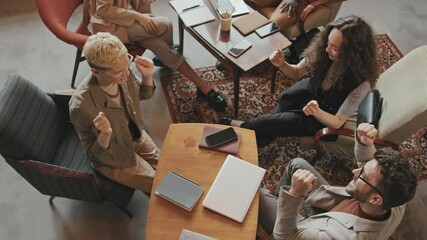 Top-view slowmo of young enthusiastic team working together on important project giving high-fives to each other sitting together at work desk in modern loft coworking office