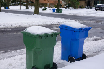 A blue and a green trash can covered with snow by the side of a snow covered, freshly plowed street...